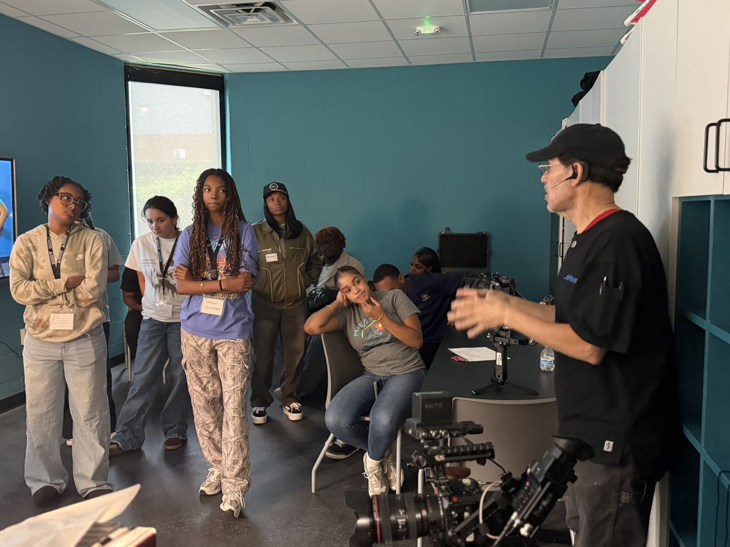 A mentor holding a camera rig while briefing students seated around tables at the NextGen Media Summit