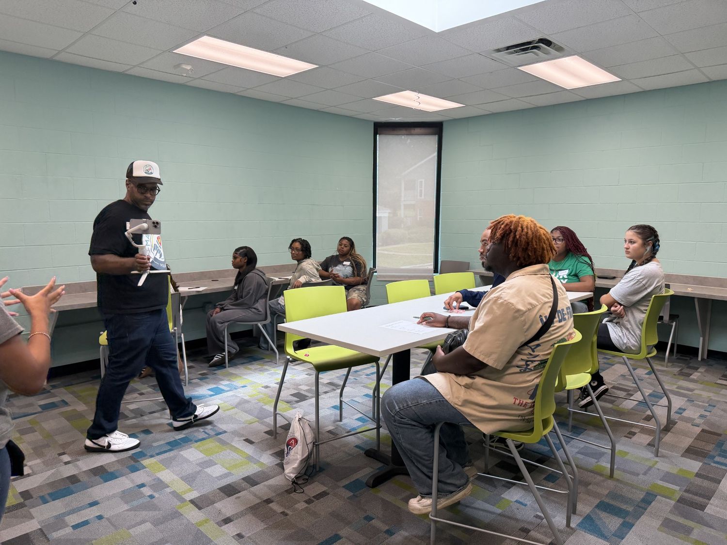 A mentor holding a camera rig while briefing students seated around tables.